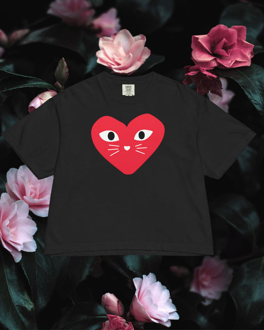 Black t-shirt with a red heart design featuring eyes and cat whiskers, placed on a floral background.