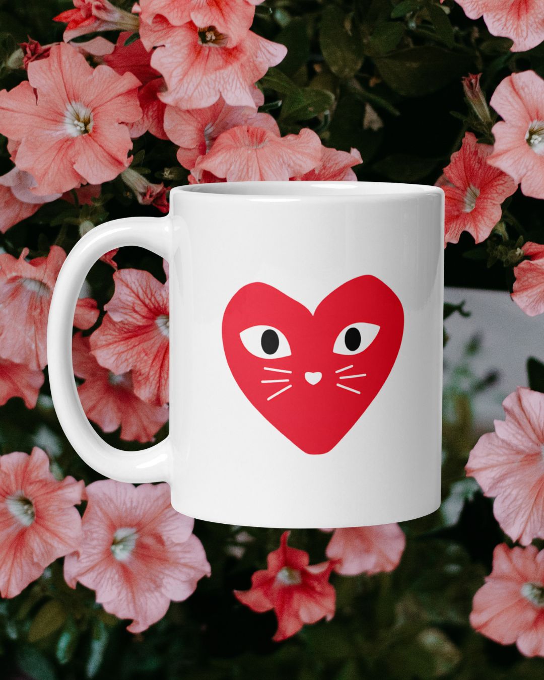 White mug with a red heart design featuring eyes and a mouth, set against a background of pink flowers.