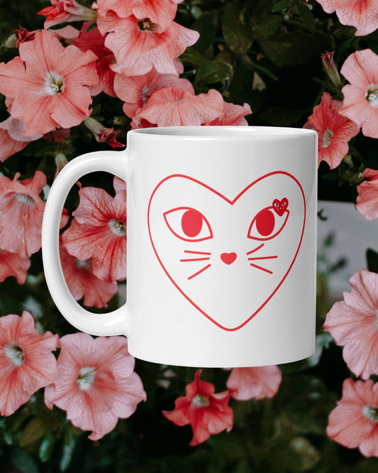 White mug with red heart-shaped cat face design against a background of pink flowers