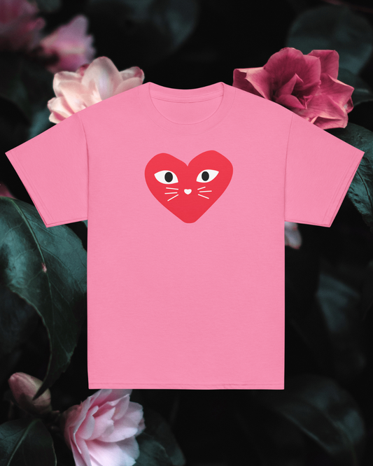 Pink t-shirt with a red heart design featuring eyes and a mouth, placed on dark green leaves with pink flowers.